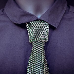 Puede incluir: Primer plano de una camisa morada oscura con cuello y una corbata de cota de malla &uacute;nica. La corbata es plateada y verde, con un patr&oacute;n texturizado en forma de escamas. El logotipo "SHAILLE CHAINMAILLE" es visible.