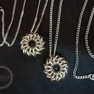 May include: Two silver chainmaille necklaces with black center pieces. The necklaces are on a black background.