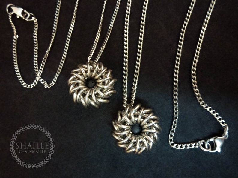 May include: Two silver chainmaille necklaces with black center pieces. The necklaces are on a black background.