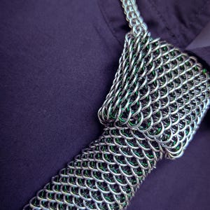Puede incluir: Primer plano de un collar de cota de malla plateada con detalles verdes. La cota de malla est&aacute; intrincadamente tejida, sobre un fondo morado oscuro. El logotipo "SHAILLE CHAINMAILLE" es visible.
