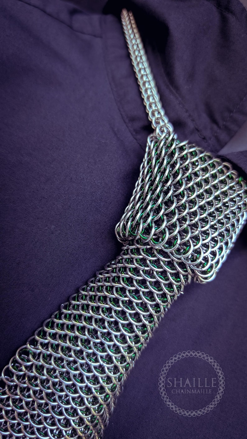 Puede incluir: Primer plano de un collar de cota de malla plateada con detalles verdes. La cota de malla est&aacute; intrincadamente tejida, sobre un fondo morado oscuro. El logotipo "SHAILLE CHAINMAILLE" es visible.