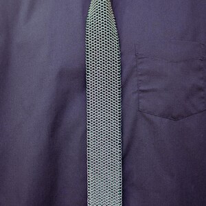 Puede incluir: Una corbata de cota de malla plateada se muestra sobre una camisa azul oscuro. La corbata est&aacute; hecha de peque&ntilde;os anillos de metal entrelazados, creando una tela texturizada y flexible. La camisa tiene un bolsillo y la marca "SHAILLE CHAINMAILLE" es visible.