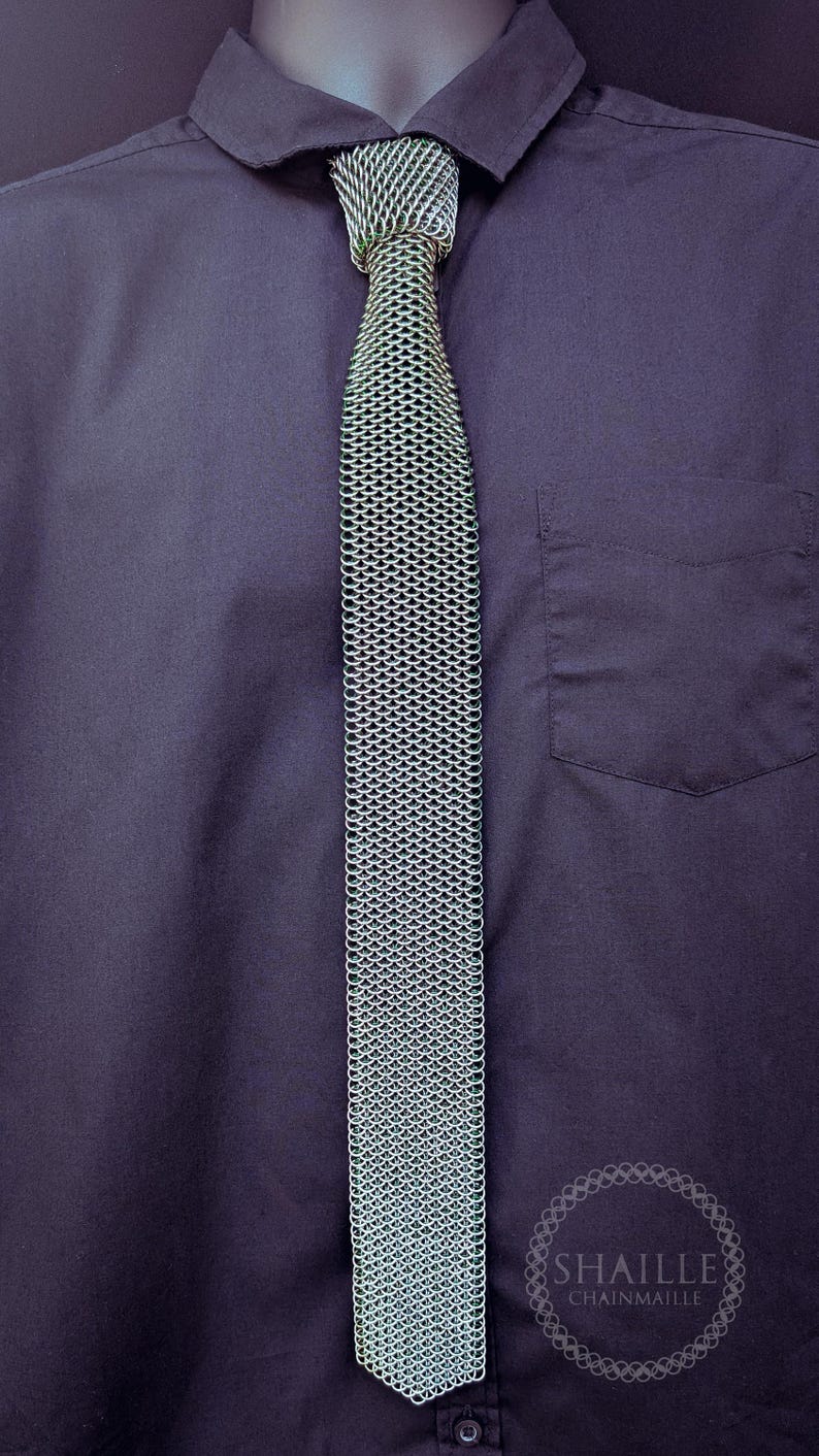 Puede incluir: Una corbata de cota de malla plateada se muestra sobre una camisa azul oscuro. La corbata est&aacute; hecha de peque&ntilde;os anillos de metal entrelazados, creando una tela texturizada y flexible. La camisa tiene un bolsillo y la marca "SHAILLE CHAINMAILLE" es visible.