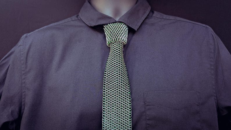 Puede incluir: Primer plano de una camisa morada oscura con una corbata verde y plateada estilo cota de malla. La corbata est&aacute; anudada y centrada, con el cuello de la camisa abierto. La camisa tiene un bolsillo en el lado izquierdo.