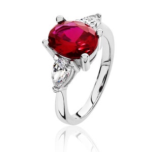 May include: A silver ring with a large, oval, ruby-red gemstone at its centre. Two pear-cut, clear gemstones are either side of the main stone. The ring's band is smooth and polished. A classic and elegant piece of jewellery.