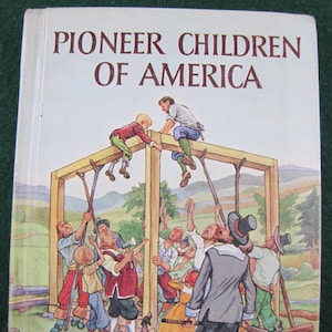 May include: A vintage book cover titled "Pioneer Children of America" featuring an illustration of people constructing a wooden structure. The cover art depicts a group of people, including children, working together, with tools and a dog visible.