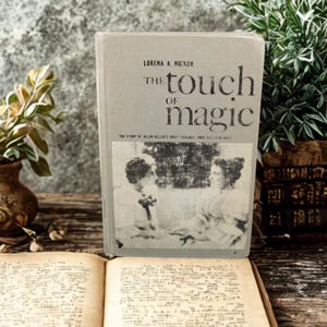 May include: A vintage book titled "The Touch of Magic" by Lorena A. Hickok. The book has a light gray cover with black text and a black and white image. An open book with handwritten text is in the foreground.
