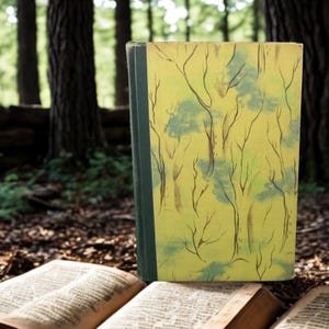 May include: A vintage book with a yellow cover featuring a painted forest scene with brown tree trunks and green foliage. The book's spine is dark green. Open books are in the foreground, with a forest backdrop.