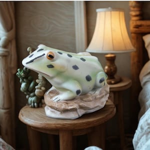 May include: A ceramic frog figurine in shades of green and white with black spots and orange eyes. The frog is perched on a textured, beige rock base. The figurine is displayed on a small wooden table.