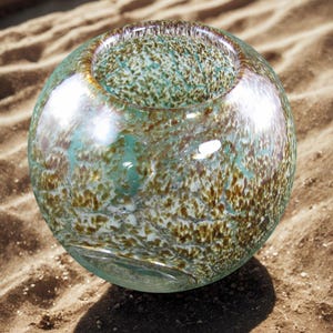 May include: A clear glass bowl with a speckled pattern in shades of teal, brown, and white. The round bowl has a wide opening and sits on a sandy surface. The glass reflects light, creating highlights and shadows.