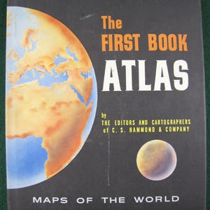 May include: A vintage atlas titled "The First Book Atlas" with a world map illustration on the cover. The cover is black with orange and white text. The atlas is published by C.S. Hammond & Company and features maps of the world.