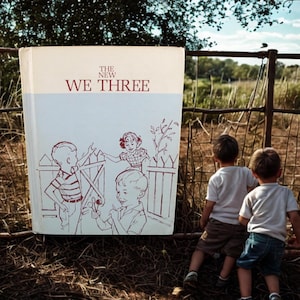 May include: A vintage hardcover book titled THE NEW WE THREE with a cream and light blue cover. The front features a maroon line drawing of three children near a picket fence. The book is positioned in front of a natural outdoor setting.