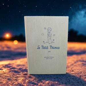 May include: A hardcover book with a cream-colored fabric cover featuring a blue line drawing of a small prince standing on a planet with stars. The text reads Le Petit Prince and Educational Edition in blue ink centered on the front cover.