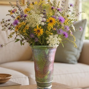 May include: A tall, flared ceramic vase with a multi-colored iridescent glaze in shades of green, pink, and purple, featuring a distinct circular base. The vase is filled with a large, dense arrangement of wildflowers, including yellow black-eyed Susans, white Queen Anne's lace, and purple lavender sprigs.