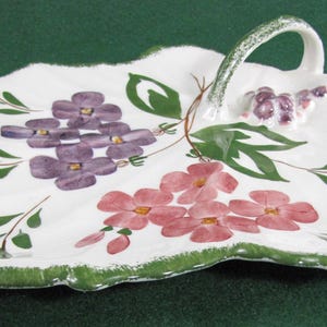 May include: A white ceramic serving dish shaped like a leaf with a handle. The dish is decorated with hand-painted purple and pink flowers and green leaves.