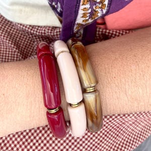 May include: A set of three beaded bracelets in shades of red, pink, and brown. The bracelets are made of cylindrical beads with gold-colored connectors. The background includes a purple scarf with a floral pattern and a red and white checkered pattern.