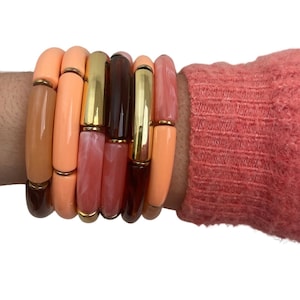 May include: A stack of colourful bracelets in shades of peach, pink, and brown with gold accents. The bracelets are made of plastic and have a smooth, glossy finish.