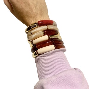May include: A stack of colorful bangle bracelets with gold accents. The bracelets are made of different materials, including resin and wood. The bracelets are stacked on a person's wrist.