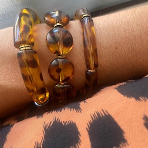 Pode incluir: Três pulseiras âmbar com detalhes dourados. As pulseiras apresentam uma mistura de contas redondas e cilíndricas. O fundo mostra um tecido estampado em tons de castanho e preto.