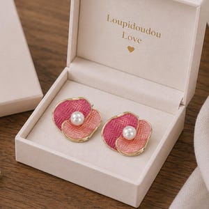 May include: A pair of flower-shaped earrings with a pearl center, presented in a white box. The earrings feature pink and red petals with a gold-colored trim. The box is labeled "Loupidoudou Love".
