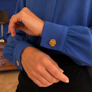 Pode incluir: Uma blusa azul de manga comprida com punhos franzidos presos por botões redondos e texturizados em tom dourado. O tecido tem um acabamento liso e os punhos ajustam-se ao pulso, destacando o detalhe metálico dos fechos circulares.