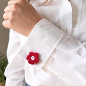 Pode incluir: Uma camisa branca de manga comprida com um acessório de flor vermelha em croché preso ao punho. A flor tem seis pétalas arredondadas e um pequeno botão branco de quatro furos fixado no centro da flor.