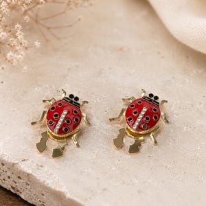 May include: Two ladybird-shaped earrings. Each earring has a red body with black spots, a black head, and gold-coloured legs. The ladybird's body is decorated with a row of clear crystals. The earrings are on a neutral background.