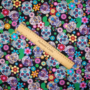 Op de afbeelding: Zwarte stof met een herhalend patroon van kleurrijke suiker schedels en bloemen. De stof is versierd met verschillende kleuren, waaronder roze, geel, blauw, groen en rood.