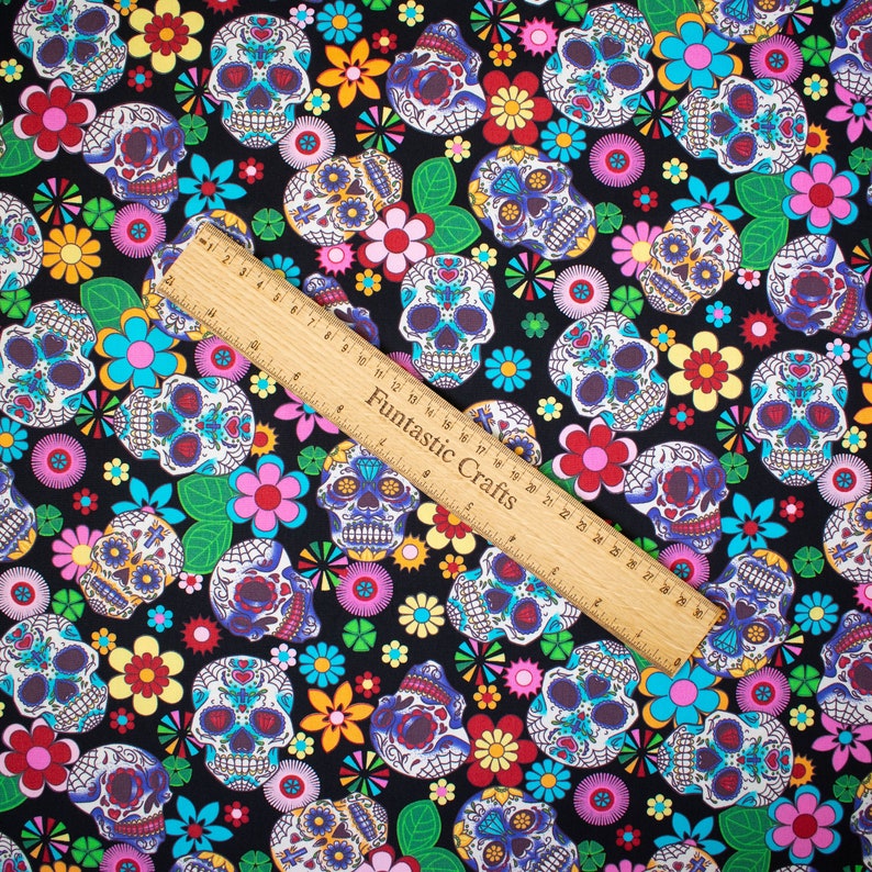 Op de afbeelding: Zwarte stof met een herhalend patroon van kleurrijke suiker schedels en bloemen. De stof is versierd met verschillende kleuren, waaronder roze, geel, blauw, groen en rood.