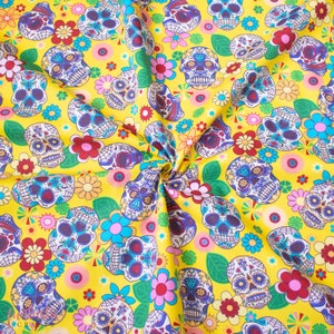 Op de afbeelding: Gele stof met een herhalend patroon van kleurrijke suiker schedels en bloemen. De schedels hebben ingewikkelde ontwerpen en zijn omringd door roze, rode en gele bloemen.