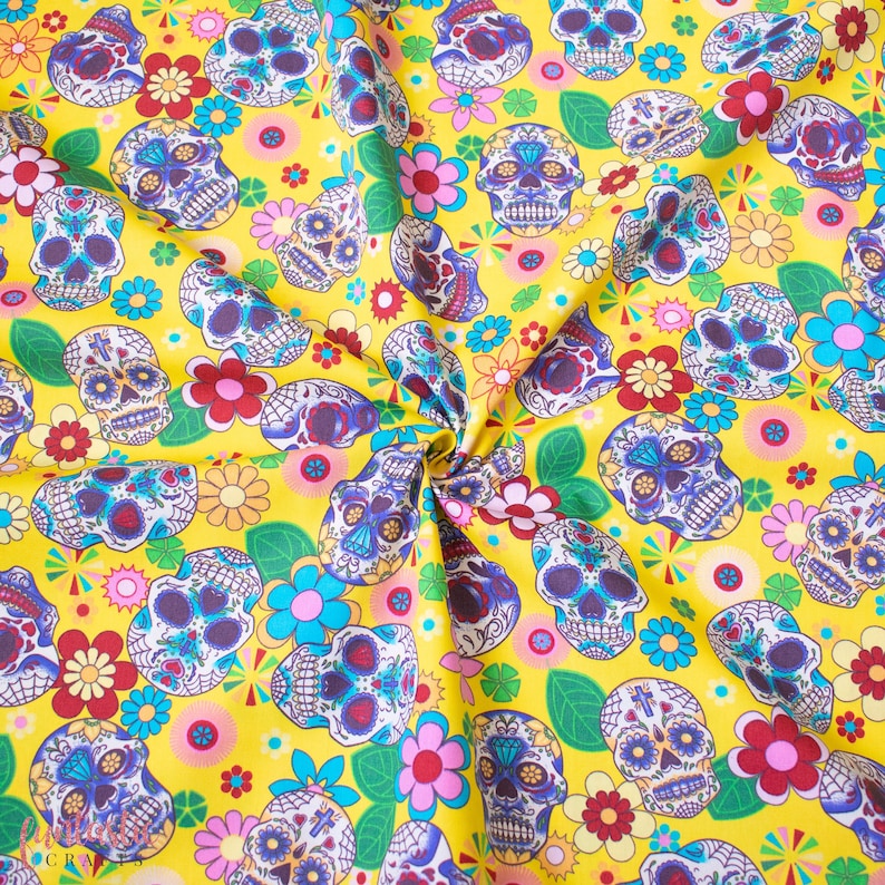 Op de afbeelding: Gele stof met een herhalend patroon van kleurrijke suiker schedels en bloemen. De schedels hebben ingewikkelde ontwerpen en zijn omringd door roze, rode en gele bloemen.
