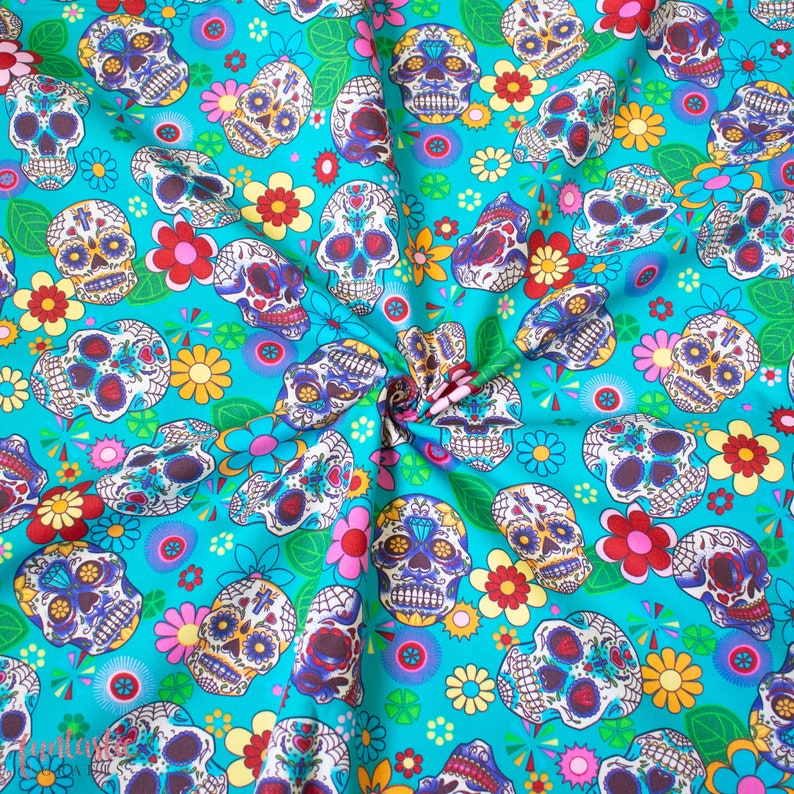 Op de afbeelding: Een turquoise stof met een herhalend patroon van kleurrijke suiker schedels en bloemen. De schedels zijn versierd met ingewikkelde details en hebben een eigenzinnige, feestelijke uitstraling.