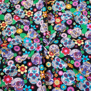 Op de afbeelding: Zwarte stof met een herhalend patroon van kleurrijke suiker schedels en bloemen. De schedels hebben ingewikkelde ontwerpen en zijn omringd door roze, gele, blauwe en groene bloemen.