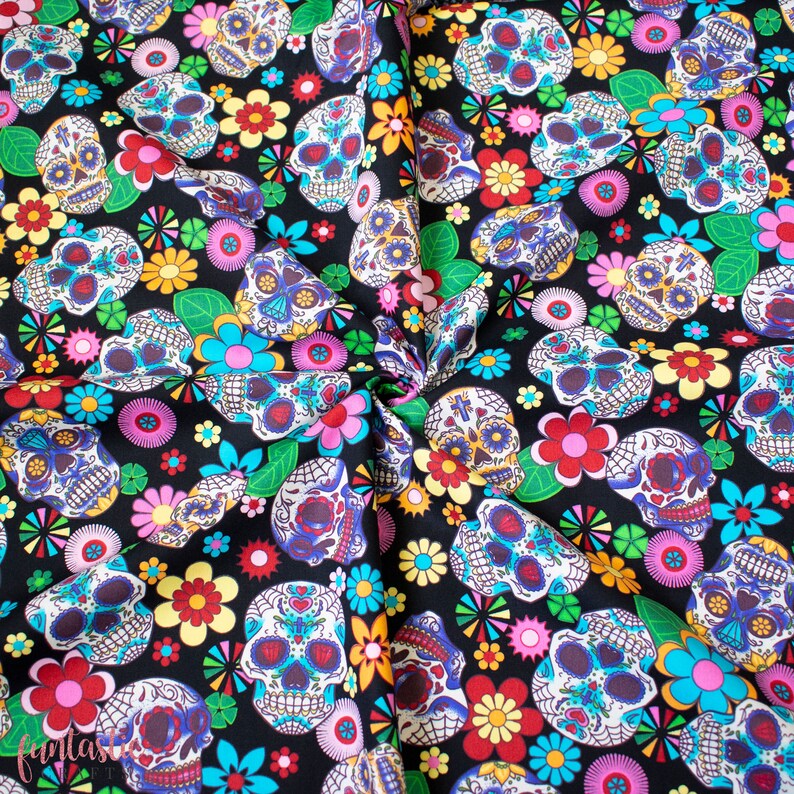 Op de afbeelding: Zwarte stof met een herhalend patroon van kleurrijke suiker schedels en bloemen. De schedels hebben ingewikkelde ontwerpen en zijn omringd door roze, gele, blauwe en groene bloemen.