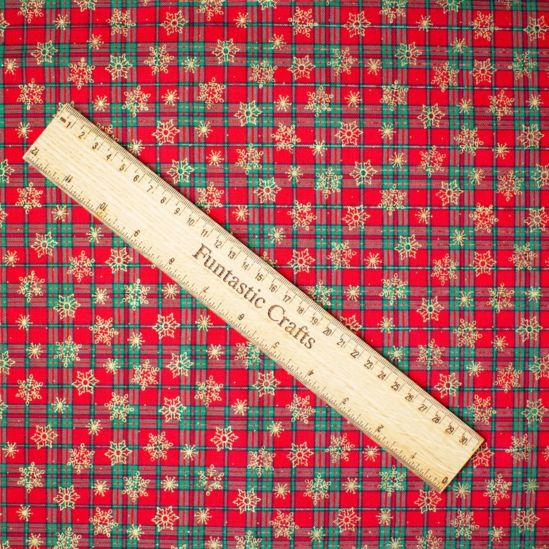 K&ouml;nnte beinhalten: Roter und gr&uuml;ner karierter Stoff mit einem goldenen Schneeflockenmuster. Ein Holzliniall mit der Aufschrift "Fantastic Crafts" liegt auf dem Stoff.