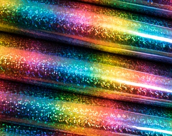 Op de afbeelding: Close-up van een regenboogkleurige glitterstof met een holografische afwerking. De stof is gelaagd en gevouwen, waardoor een textuur en een driedimensionaal effect ontstaat.
