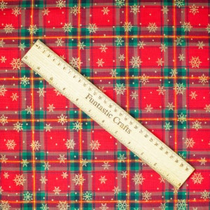 K&ouml;nnte beinhalten: Roter und gr&uuml;ner karierter Stoff mit goldenem Schneeflockenmuster. Ein Holzliniall mit der Aufschrift "Fantastic Crafts" liegt auf dem Stoff.