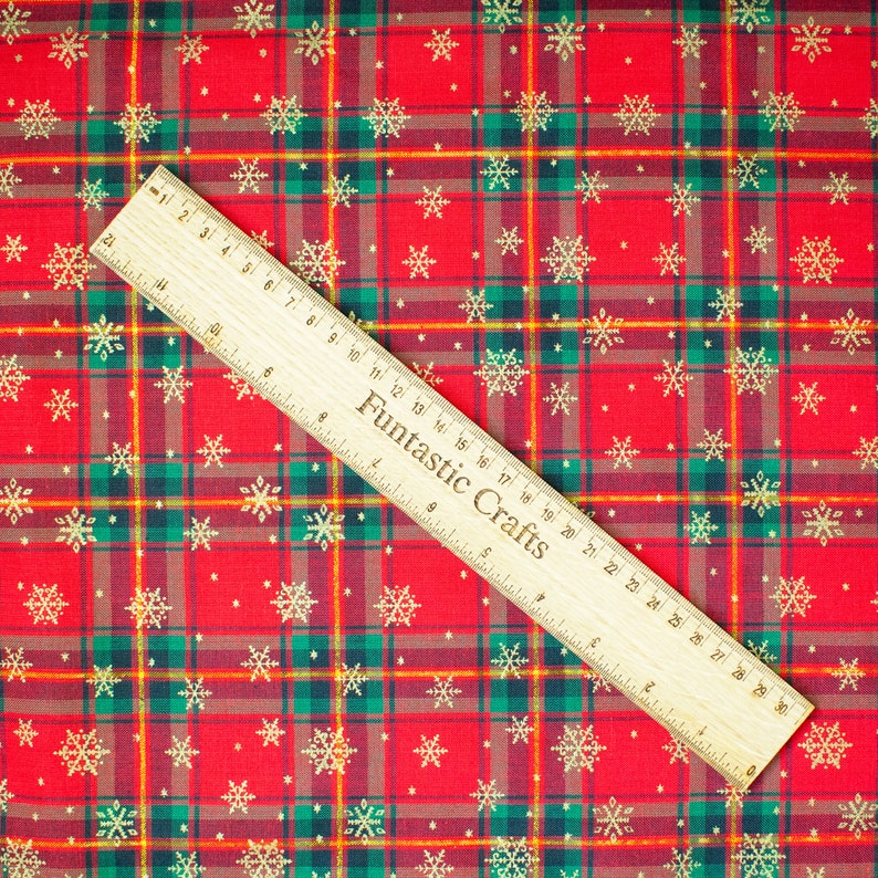 K&ouml;nnte beinhalten: Roter und gr&uuml;ner karierter Stoff mit goldenem Schneeflockenmuster. Ein Holzliniall mit der Aufschrift "Fantastic Crafts" liegt auf dem Stoff.