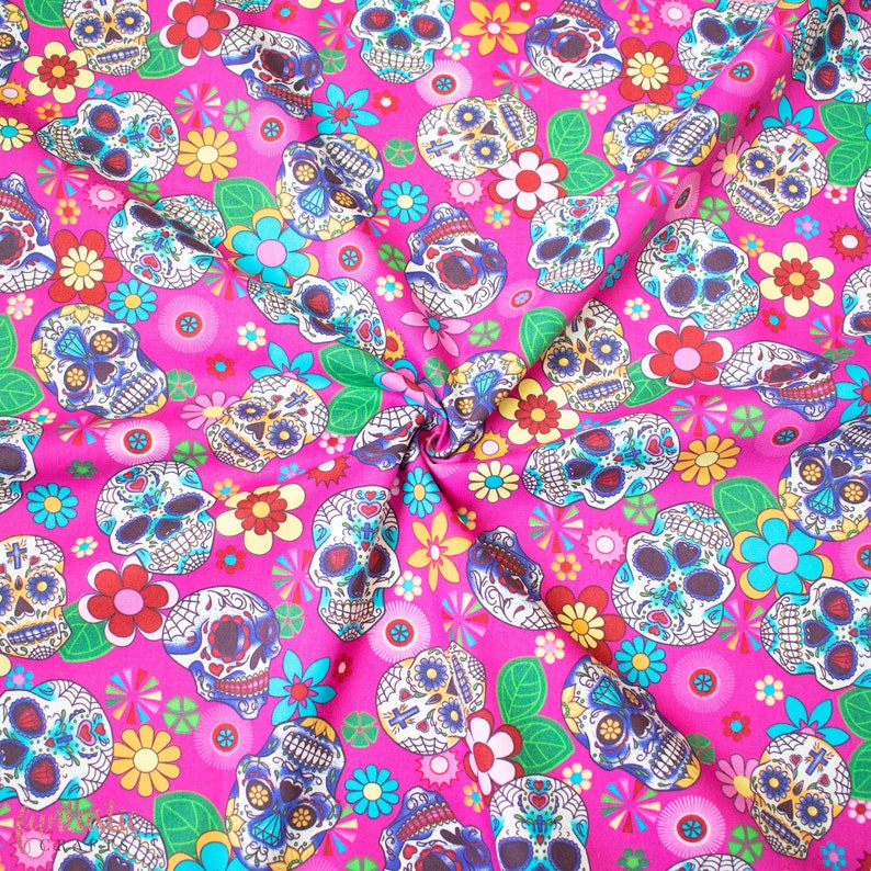 Op de afbeelding: Roze stof met een herhalend patroon van kleurrijke suiker schedels en bloemen. De schedels hebben ingewikkelde ontwerpen en zijn omringd door fel roze, gele en blauwe bloemen.
