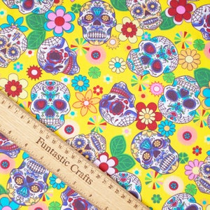 Op de afbeelding: Gele stof met een herhalend patroon van kleurrijke suiker schedels, bloemen en geometrische vormen. De suiker schedels zijn wit met zwarte contouren en hebben kleurrijke details.