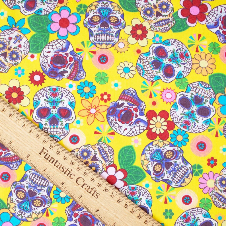 Op de afbeelding: Gele stof met een herhalend patroon van kleurrijke suiker schedels, bloemen en geometrische vormen. De suiker schedels zijn wit met zwarte contouren en hebben kleurrijke details.
