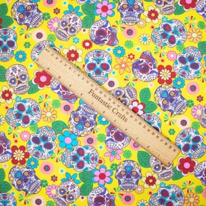Op de afbeelding: Gele stof met een herhalend patroon van kleurrijke suiker schedels en bloemen. De stof wordt gemeten met een houten liniaal waarop "Funfantastic Crafts" staat.