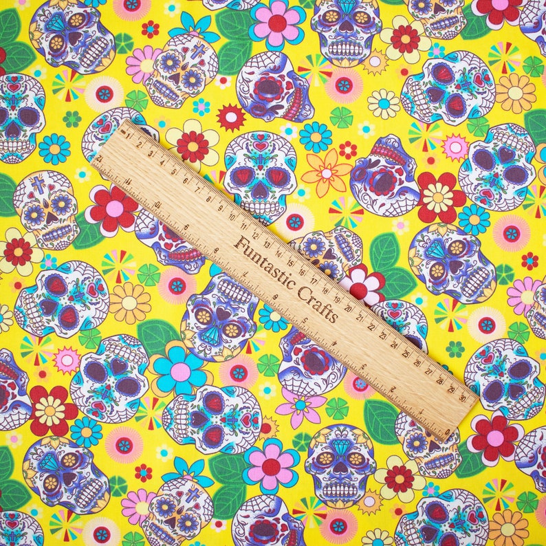 Op de afbeelding: Gele stof met een herhalend patroon van kleurrijke suiker schedels en bloemen. De stof wordt gemeten met een houten liniaal waarop "Funfantastic Crafts" staat.