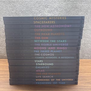 Könnte beinhalten: Ein vertikaler Stapel von zwanzig dunkelgrauen Hardcover-Büchern aus der Reihe Voyage Through the Universe. Jeder Buchrücken zeigt den Serientitel und die einzelnen Buchtitel in verschiedenen Farben, darunter Cosmic Mysteries, Spacefarers und Frontiers of Time.