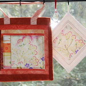 May include: A square fabric tote bag in shades of rust and orange with a central panel featuring a floral print, paired with a diamond-shaped white fabric potholder displaying a matching floral pattern with decorative stitching and a hanging loop.