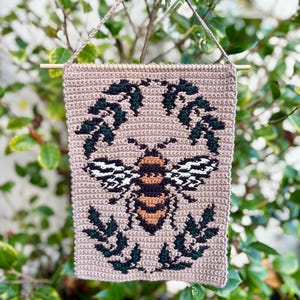 May include: A beige crochet wall hanging with a bee motif. The bee is detailed with black, white, and orange. It is encircled by a wreath of green leaves. The hanging is suspended by a wooden dowel and a beige crochet chain.