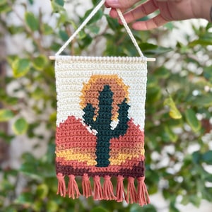 Puede incluir: Un tapiz de pared rectangular hecho a mano con un diseño de paisaje desértico. La pieza de arte de ganchillo representa un cactus verde, una puesta de sol amarilla y naranja y montañas de tonos rojos. El borde inferior tiene borlas naranjas a juego.