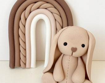 May include: A brown and beige fondant bunny figurine with a stitched heart on its chest, sitting in front of a brown, beige, and white fondant rainbow.