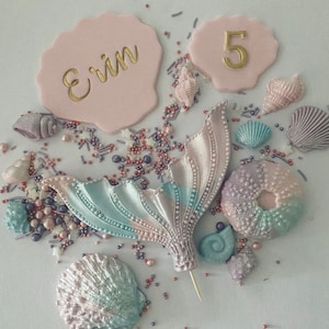 May include: A pink cake topper with the name "Erin" and the number "5" in gold lettering. The topper is surrounded by pastel blue, pink, and purple seashells and sprinkles.