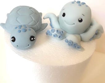 May include: Two light blue fondant sea creatures: a turtle and an octopus. The turtle has a patterned shell and a smiling face. The octopus has eight tentacles and a smiling face. Both have black eyes and blue accents.